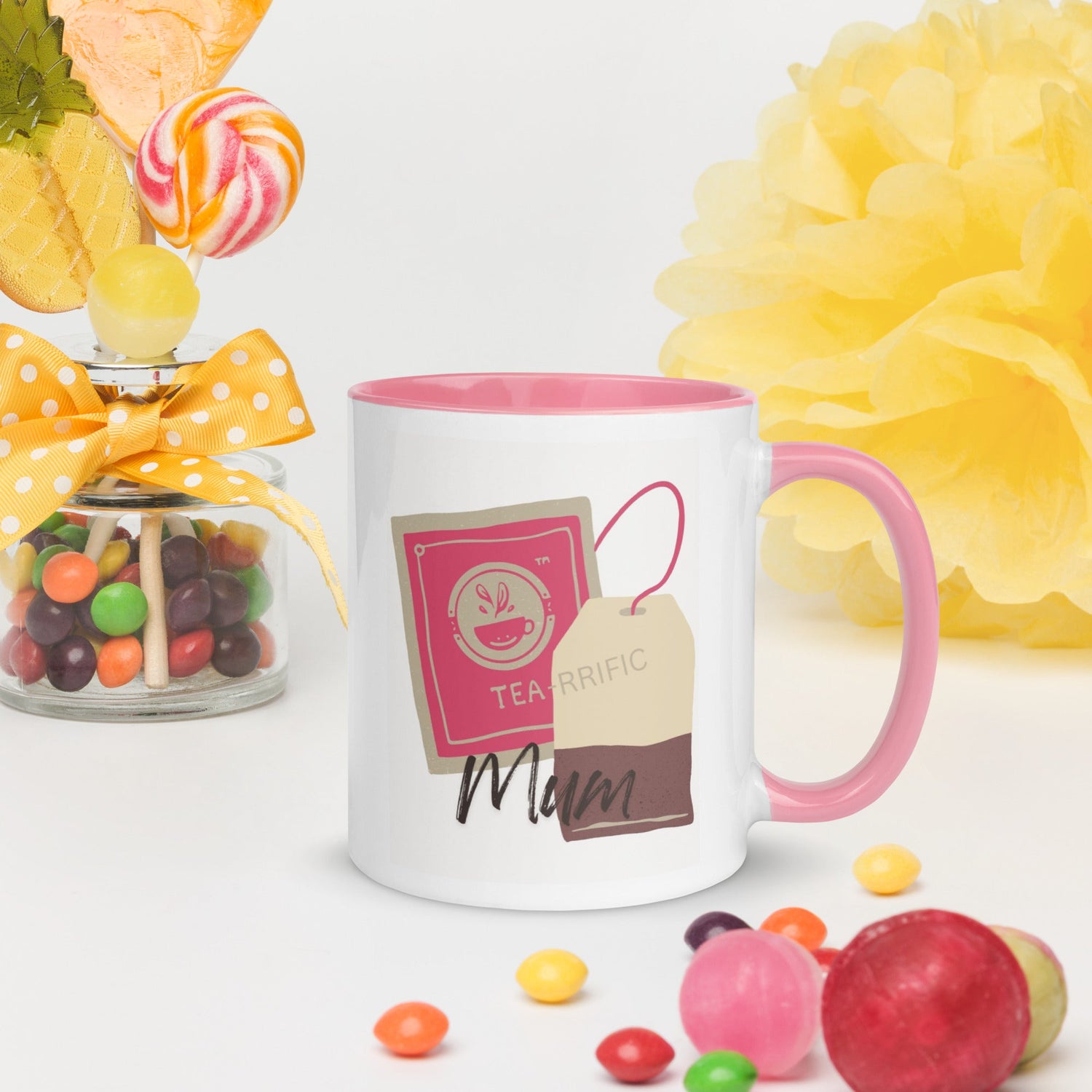 White ceramic mug with pink handle and interior, featuring a 'Terrific Mum' tag, surrounded by colourful candies and flowers.